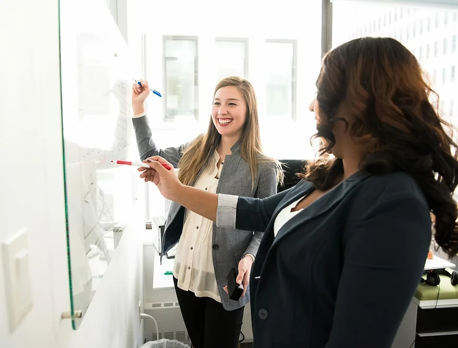 Twee vrouwelijke collega’s schrijven samen op een whiteboard tijdens een brainstormsessie op kantoor.