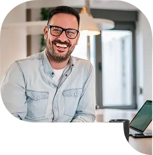 Lachende man met bril zit aan een bureau met een laptop en koffiemok in een moderne werkomgeving.