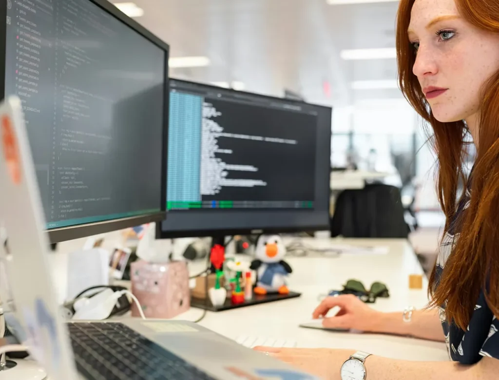 Vrouw werkt op kantoor met programmeercode op twee beeldschermen.