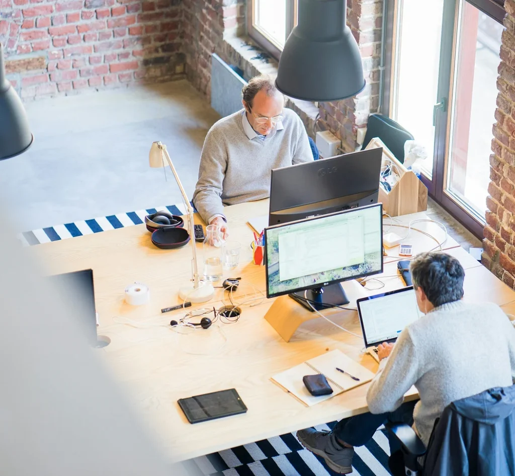Twee collega’s werken op laptops aan een gedeeld bureau in een modern kantoor, van bovenaf gezien.