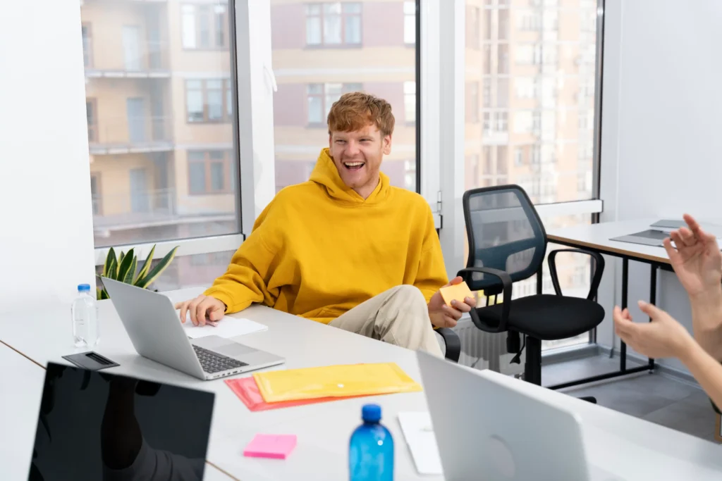 lachende man in gele hoodie die achter zijn bureau zit met een sticky note blok in zijn handen